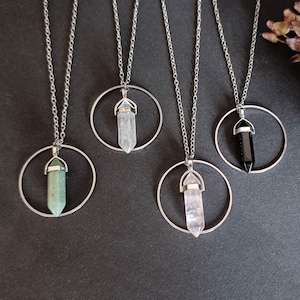 Può includere: Quattro collane con ciondoli di cristallo con catene d'argento e montature circolari. I cristalli sono nelle tonalità di verde, trasparente, rosa e nero. Le collane sono esposte su una superficie scura.