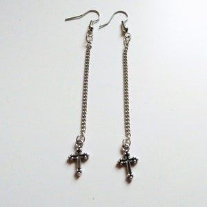 Op de afbeelding: Een paar zilveren oorbellen met een lange ketting en een klein kruisje aan het einde.