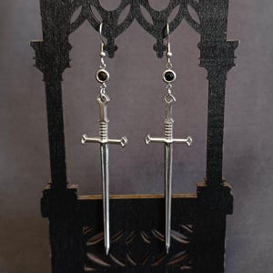 Può includere: Un paio di orecchini a spada color argento con dettagli di pietre preziose nere. Gli orecchini sono esposti su un supporto decorativo nero su sfondo scuro. Le spade hanno elsa e lame dettagliate.
