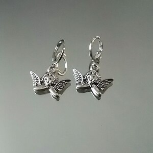 May include: Silver hoop earrings with a small angel charm hanging from each hoop.