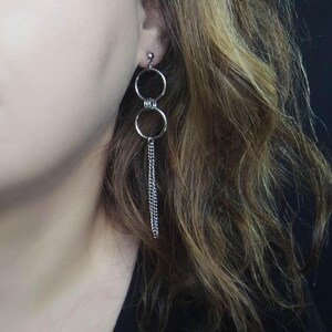 May include: Silver-toned dangle earrings. Each earring features two connected circular hoops, one above the other, with a delicate chain hanging from the bottom hoop. The earrings are shown against a dark background.