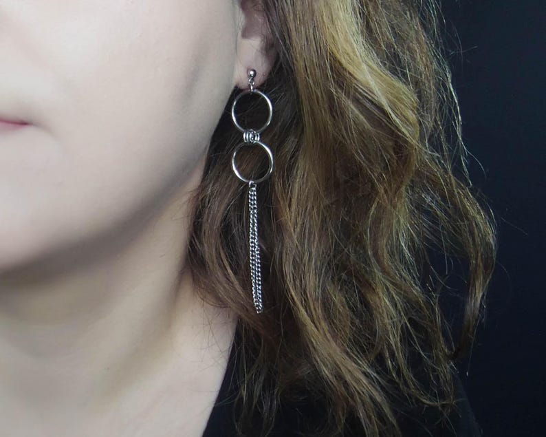 May include: Silver-toned dangle earrings. Each earring features two connected circular hoops, one above the other, with a delicate chain hanging from the bottom hoop. The earrings are shown against a dark background.