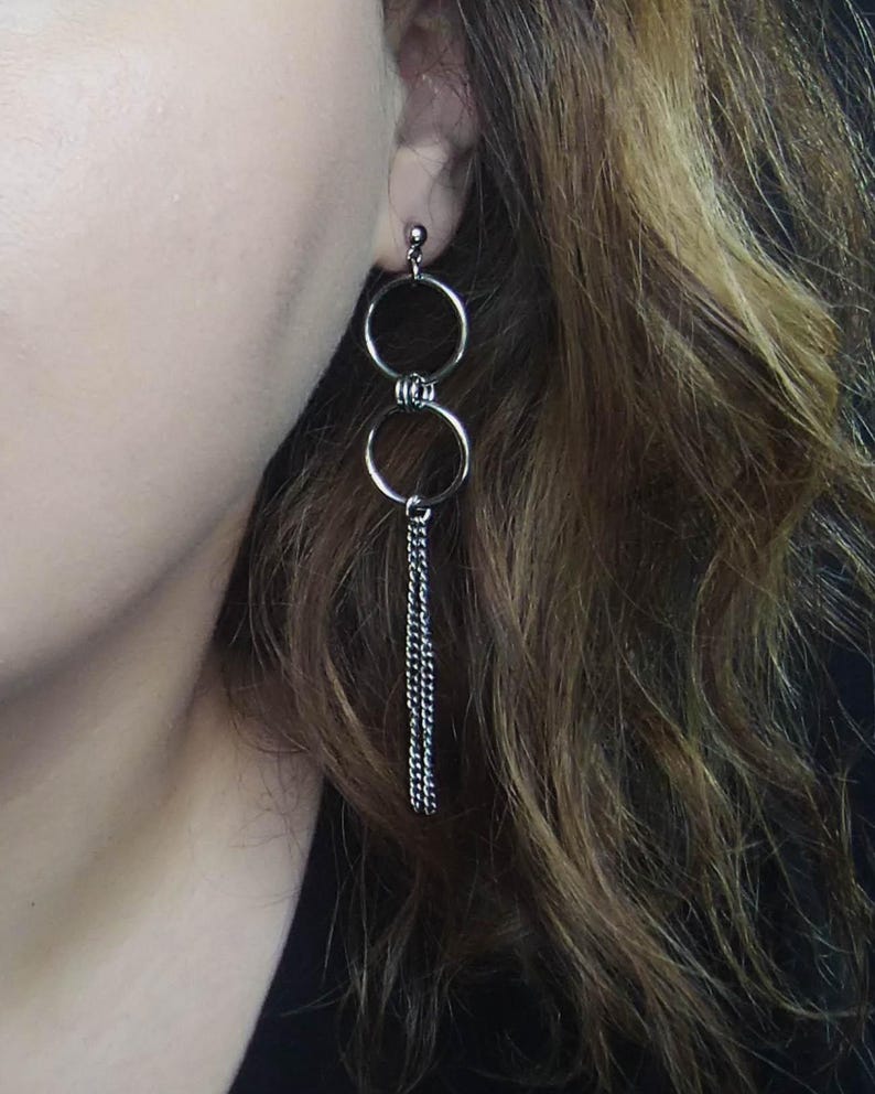 May include: Silver-toned dangle earrings featuring two connected circular hoops. A delicate chain of silver links hangs from the bottom hoop. The earrings are shown against a background of wavy brown hair.