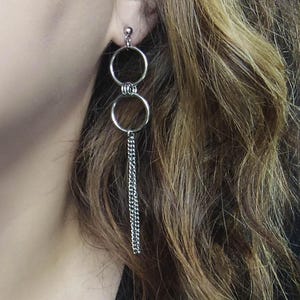May include: Silver-toned drop earrings featuring two connected circular hoops, with a delicate chain dangling from the lower hoop. The earrings are shown against a backdrop of wavy, light brown hair.