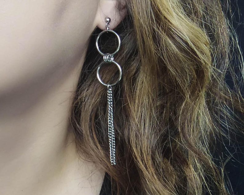 May include: Silver-toned drop earrings featuring two connected circular hoops, with a delicate chain dangling from the lower hoop. The earrings are shown against a backdrop of wavy, light brown hair.