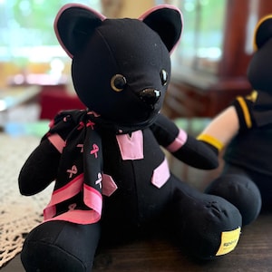 May include: A black plush bear featuring pink trim on the ears, a pink V-neck accent on the chest, and a black scarf patterned with pink ribbons. The bear has dark button eyes, a black nose, and a small yellow tag on the foot labeled spirit.
