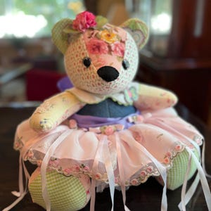 May include: A stuffed bear toy crafted from various floral and green gingham patterned fabrics. The bear features a pink fabric flower on its head, black button eyes, and a dark nose. It wears a purple waistband and a layered pink tulle skirt.