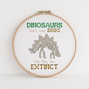 May include: A white embroidery hoop with a cross-stitch design featuring a dinosaur skeleton and the text "Dinosaurs didn't read books, now they are extinct".