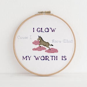 May include: A circular wooden embroidery hoop containing white fabric with a cross-stitch design. The design features a brown unicorn jumping over pink clouds, with the text I GLOW, Cause I, Know What, MY WORTH IS stitched in blue and pink.