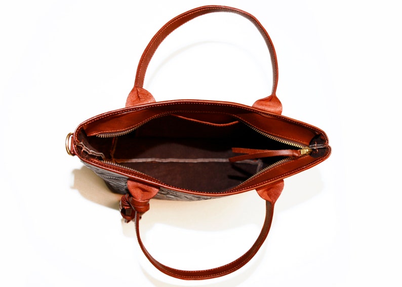Op de afbeelding: Een bruine leren tote bag met een ritssluiting en een bovenste handvat. De tas is open en de binnenkant is gevoerd met een bruine stof.