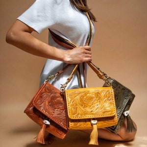 May include: Three leather crossbody bags with intricate floral designs. The bags are in shades of brown, yellow, and green. Each bag has a tassel hanging from the bottom.