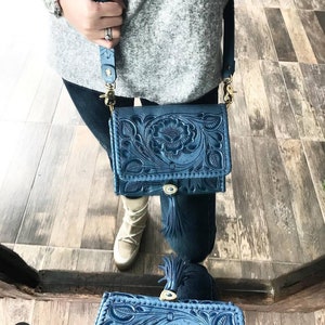 May include: A blue leather crossbody bag with intricate floral tooling and a tassel. The bag has a gold-tone buckle and strap.