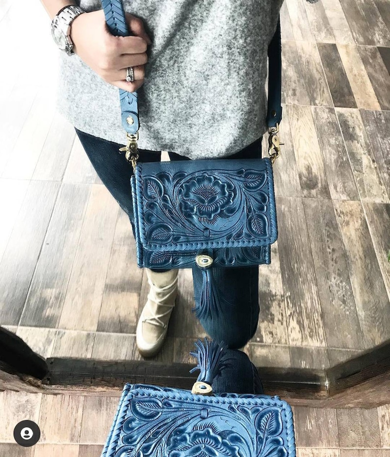 May include: A blue leather crossbody bag with intricate floral tooling and a tassel. The bag has a gold-tone buckle and strap.