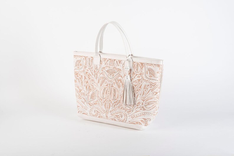 May include: A white leather tote bag with a floral design. The bag has a top handle and a tassel.