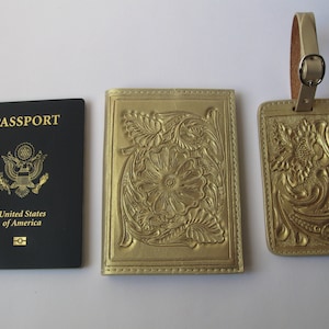 May include: A gold passport holder and luggage tag with a floral embossed design. The passport holder has the word "PASSPORT" and "United States of America" printed on it.