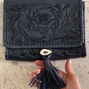 May include: A black leather clutch with a floral embossed design and a tassel. The clutch has a braided trim and a small button closure.