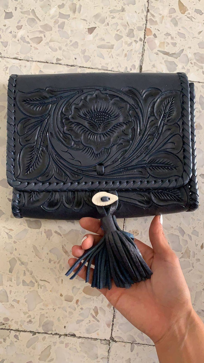 May include: A black leather clutch with a floral embossed design and a tassel. The clutch has a braided trim and a small button closure.