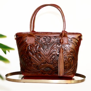 Op de afbeelding: Een bruine leren tote bag met een reli&euml;f van een bloemenpatroon en een kwast. De tas heeft een bovenste handvat en een schouderband.