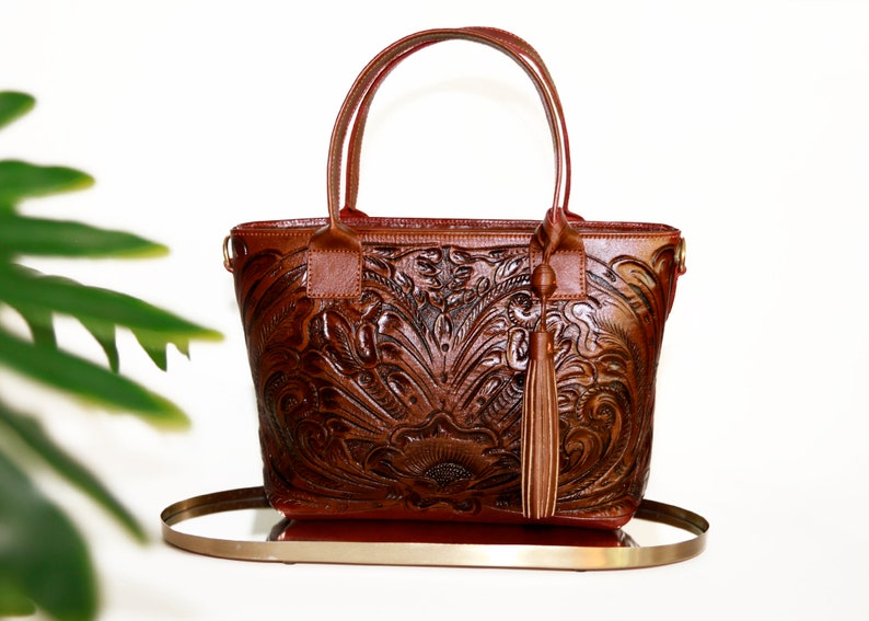 Op de afbeelding: Een bruine leren tote bag met een reli&euml;f van een bloemenpatroon en een kwast. De tas heeft een bovenste handvat en een schouderband.