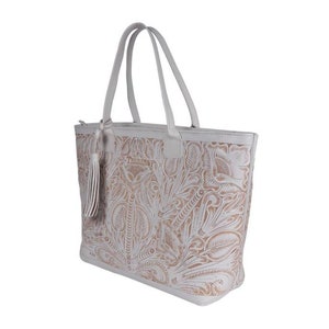 May include: White leather tote bag with a floral embossed design. The bag has a zipper closure and a tassel on the front. The brand name "The Sak" is embossed on the front of the bag.