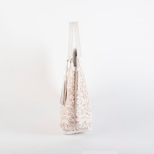 May include: A white leather tote bag with an embossed floral design and a tassel. The bag has a top handle and a zipper closure.