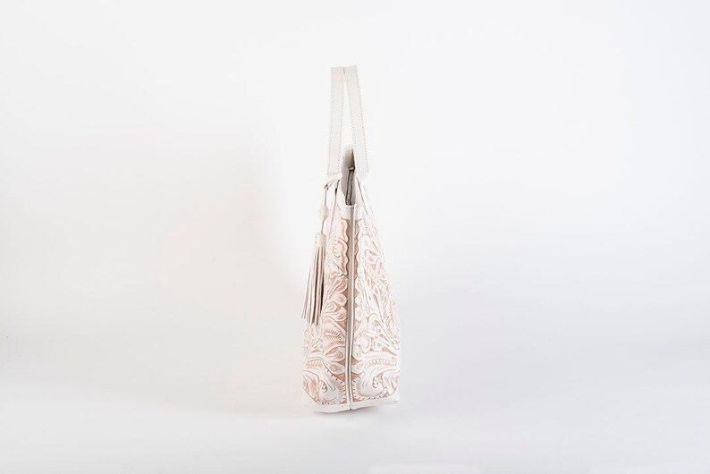 May include: A white leather tote bag with an embossed floral design and a tassel. The bag has a top handle and a zipper closure.
