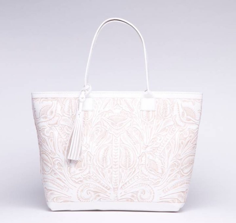 May include: White leather tote bag with an embossed floral design. The bag has a top zipper closure and a tassel.