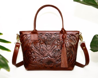 May include: A brown leather tote bag with an intricate floral embossed design. The bag has a top handle and a long adjustable shoulder strap. It features a tassel on the front.