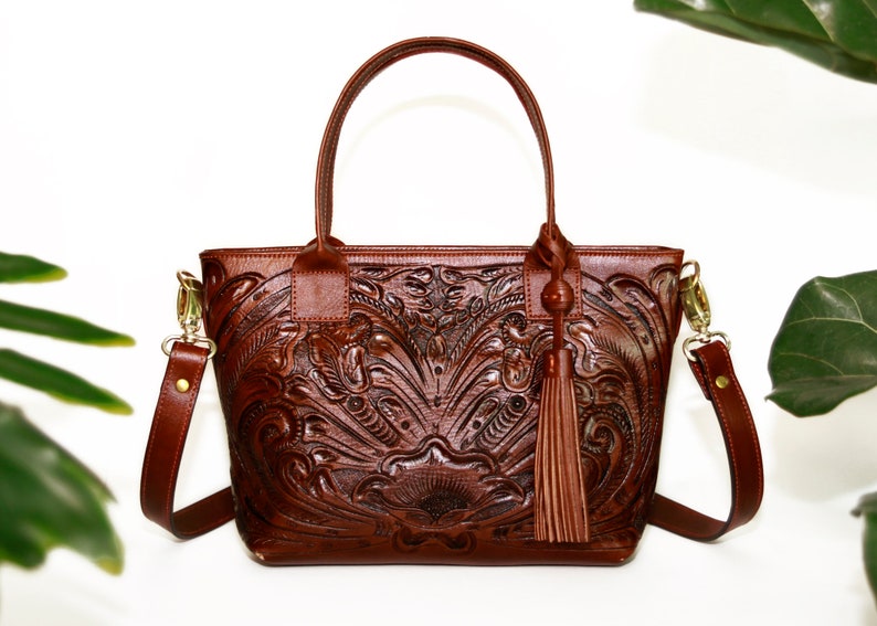Op de afbeelding: Een bruine leren tote bag met een ingewikkeld gegraveerd bloemenpatroon. De tas heeft een bovenste handvat en een lange verstelbare schouderband. Hij heeft een kwast aan de voorkant.