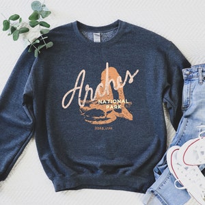 May include: A dark blue sweatshirt with a graphic print of the Arches National Park logo. The logo is in a light orange color and says "Arches National Park Moab, Utah".