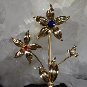 Könnte beinhalten: Eine goldfarbene Brosche mit zwei stilisierten Blumen mit blauen und roten Edelsteinen und einem Perlenakzent. Die Blumen haben zarte, strukturierte Blütenblätter und sind durch einen Stiel mit Blättern verbunden.