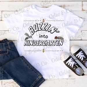 Puede incluir: Camiseta blanca con un gráfico en blanco y negro que dice "Buzzin' into Kindergarten". El gráfico incluye abejas y moscas. La camiseta está sobre una superficie de madera blanca con un par de zapatillas negras.