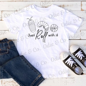 May include: White t-shirt with a black graphic of three pill bugs and the text "Just Roll with it". The shirt is on a white wooden surface with a pair of blue jeans and black sneakers.