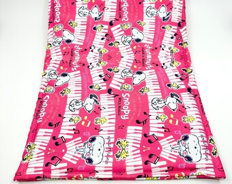 May include: Pink and white fabric cushion with a repeating pattern of Snoopy playing the piano with musical notes and the text "Snoopy with Music" and "atunanuts jazz n' music".