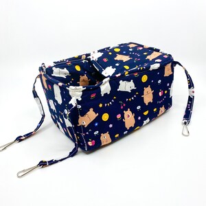 May include: A blue fabric hanging toy for small animals, featuring a white and brown bear print with fruit and party flag designs. The toy has two straps with metal clips.