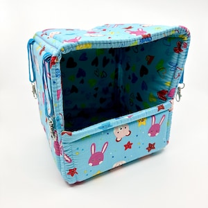 Puede incluir: Un transportín para mascotas azul claro con un estampado de conejitos rosas, estrellas y corazones. El transportín rectangular tiene una abertura con cremallera y correas azules con clips metálicos. El interior también tiene un estampado de corazones.