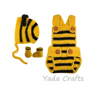 May include: A yellow and black crochet baby outfit set, including a hat, booties, and a romper. The set features a striped pattern and wooden buttons. The outfit is designed to resemble a bee.