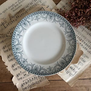 May include: A round white ceramic plate featuring a decorative border with a dark grey intricate floral and scrollwork pattern. The rim of the plate is adorned with a repeating geometric chequered design that frames the detailed botanical motifs.