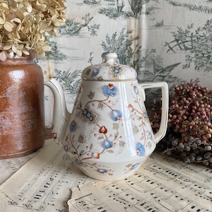 Peut inclure: Une théière en céramique de couleur crème ornée d'un motif floral délicat avec des vignes bleues, orange et vertes. La théière possède un couvercle assorti avec un petit bouton, un bec verseur courbé et une anse robuste, le tout souligné d'un fin liseré doré.