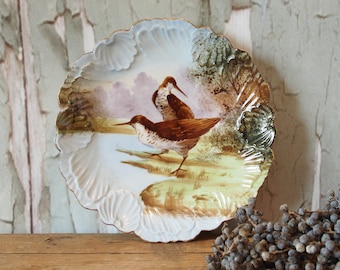 Pode incluir: Um prato de porcelana branca com borda festonada apresenta uma cena pintada à mão de dois pássaros marrons em um ambiente de floresta verde e marrom. O prato tem uma borda dourada.