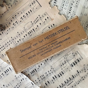 Pode incluir: Uma caixa de cartão marrom vintage com o texto "Stempel van het METRIEK-STELSEL" e "Fabrikaat en Uitgave: NOORD NEDERLANDSCHE STEMPELFABRIEK GRONINGEN (Holland)". A caixa está em cima de uma folha de música.