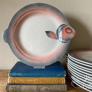 Puede incluir: Un plato de cerámica vintage con forma de pez con un diseño rosa y gris. El plato está sobre una pila de libros con el texto "B. de Spinoza ETHICA" visible.