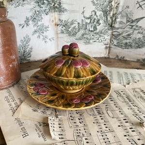 May include: A ceramic lidded dish and saucer set. The dish and saucer are decorated with a leaf and cherry design in shades of green, brown, and pink. The lid features two cherry-shaped knobs.
