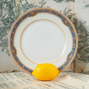 Op de afbeelding: Een witte keramische plaat met een decoratieve rand met blauwe, oranje en gouden bloempatronen. Een heldergele citroen rust voor de plaat. De achtergrond omvat vintage bladmuziek en een toile-stof.