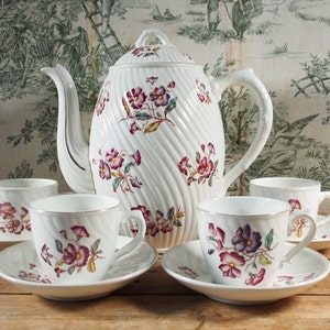 May include: A white porcelain tea set with a teapot and four teacups and saucers. The set features a floral design with purple and pink flowers. The teapot has a lid and a curved handle. The teacups have a gold rim.