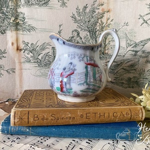 Könnte beinhalten: Eine weiße Porzellan-Milchkanne mit einem blauen und roten Blumendesign. Die Kanne hat einen abgebrochenen Rand und steht auf zwei Büchern. Das obere Buch trägt den Titel "ETHICA" auf dem Rücken.