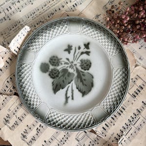 Może przedstawiać: Okrągły talerz ceramiczny z centralną, ciemnozieloną ilustracją botaniczną przedstawiającą jeżyny i liście. Brzeg ozdobiony jest wypukłym, teksturowanym wzorem kratki w odcieniach ciemnej zieleni i bieli, tworząc wyraźną obwódkę.