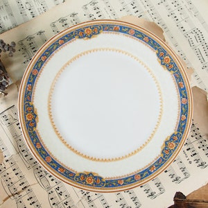 Op de afbeelding: Een witte keramische plaat met een decoratieve rand met blauwe, oranje en gouden bloempatronen. De plaat staat tegen een achtergrond van vintage bladmuziek en een houten oppervlak, wat een klassieke esthetiek cre&euml;ert.