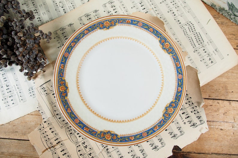 Op de afbeelding: Een witte keramische plaat met een decoratieve rand met blauwe, oranje en gouden bloempatronen. De plaat staat tegen een achtergrond van vintage bladmuziek en een houten oppervlak, wat een klassieke esthetiek cre&euml;ert.