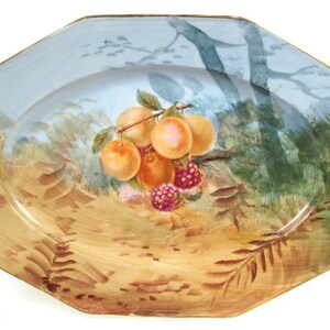 May include: A blue and white oval serving platter with a gold rim. The platter features a painted scene of a branch of fruit with peaches and raspberries, set against a background of trees and foliage.